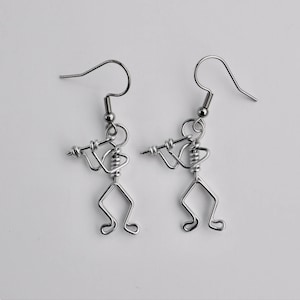 May include: A pair of silver earrings featuring a small, stylized figure playing a flute. The earrings are made of wire and have a simple, minimalist design.