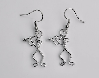 Peut inclure: Une paire de boucles d'oreilles en argent représentant une petite figurine stylisée jouant de la flûte. Les boucles d'oreilles sont en fil de fer et ont un design simple et minimaliste.