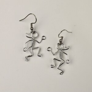 May include: A pair of silver-colored wire earrings. Each earring is shaped like a stick figure with a head, arms, legs, and a coiled spring for a torso. The earrings have a hook for pierced ears.