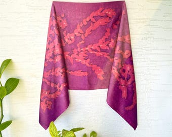 May include: A rectangular scarf in a deep magenta purple hue featuring a repeating botanical pattern of lighter pink, leaf-like shapes. The fabric has a subtle, textured sheen and hangs in a draped, folded manner against a white wall.