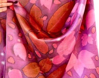 May include: A lightweight fabric scarf featuring a vibrant botanical print with overlapping leaf patterns in shades of magenta, deep purple, and burnt orange. The textile displays a smooth, semi-sheer texture with a repeating natural motif.