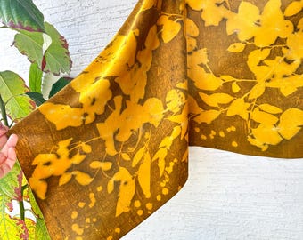 May include: A rectangular fabric scarf featuring a deep brown background with a vibrant yellow botanical print of leaves and branches. The textile displays a natural leaf-patterned design across its surface, showcasing a contrast between the dark earth-toned base and the bright yellow foliage silhouettes.