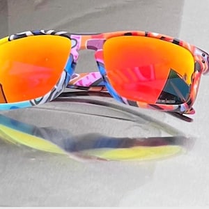 May include: Sunglasses with a multi-colored frame featuring a vibrant abstract pattern of yellow, blue, pink, and black. The lenses are reflective with a bright orange and red gradient finish, providing a bold look with a rectangular frame shape.