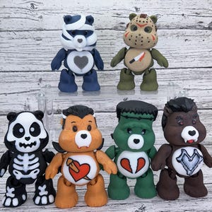 Op de afbeelding: Een verzameling van zeven kleurrijke, gestileerde berenbeeldjes. Elke beer heeft een uniek ontwerp, met thema's als een panda, Jason Voorhees, Frankenstein en een vampier. De beren staan rechtop en tonen hun kenmerkende kenmerken en outfits.