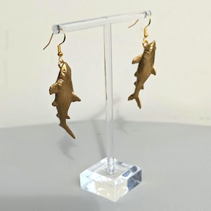 May include: Gold shark earrings hanging from a clear acrylic earring stand. The shark earrings are detailed and realistic.