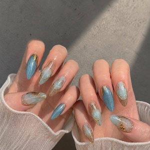Hand wearing blue marble cat eye press-on nails with gold foil accents, glossy gel finish, and long almond shape.