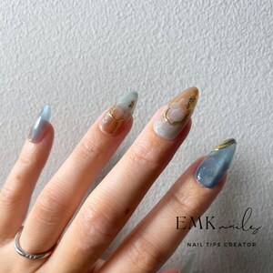 Hand wearing blue marble cat eye press-on nails with gold foil accents, glossy gel finish, and long almond shape.