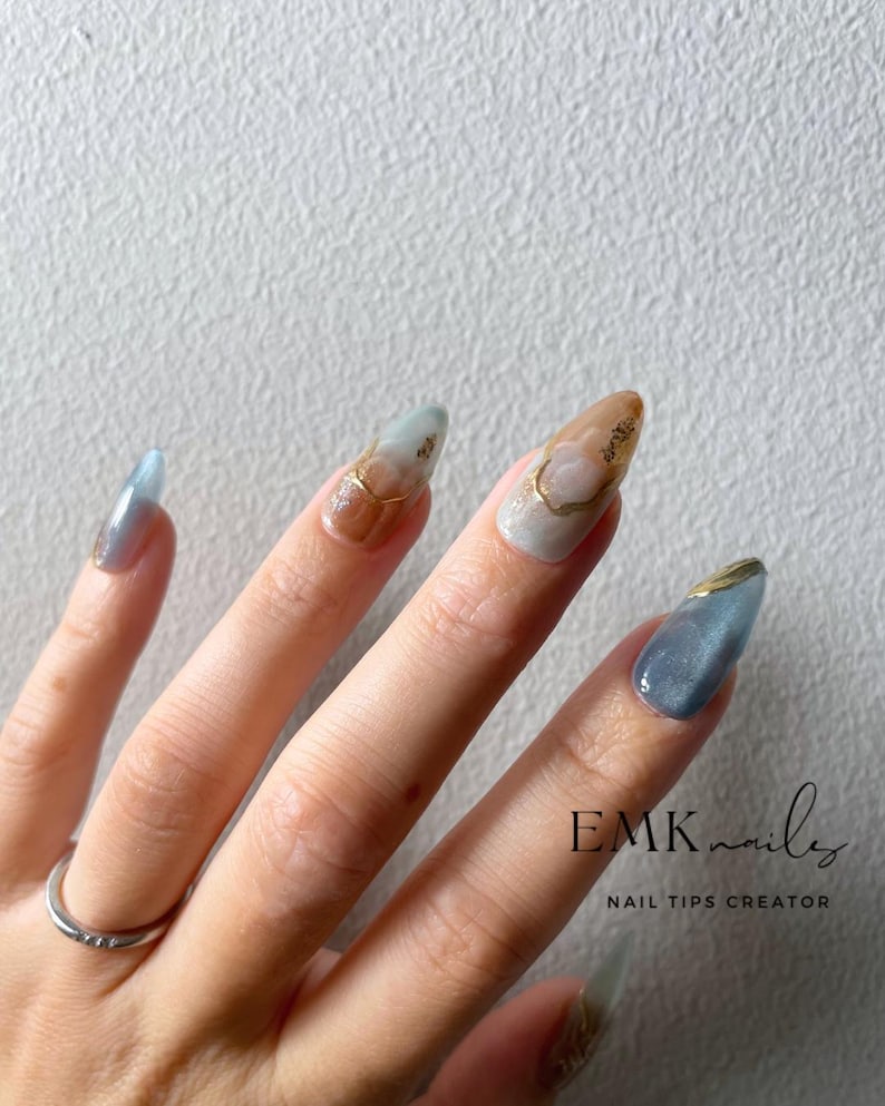 Hand wearing blue marble cat eye press-on nails with gold foil accents, glossy gel finish, and long almond shape.
