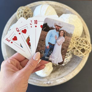 May include: A hand holds playing cards, including the King, Queen, and Ten of Hearts, and a custom card with a couple's photo. The custom card has the text "The Bishops EST. 2026". The cards are in front of a wooden bowl with decorative spheres.