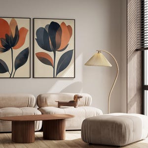 May include: Two framed abstract floral prints with orange, brown, and blue colors hang on a white wall above a beige sofa with a wooden coffee table and a beige ottoman. A floor lamp with a beige shade is in the corner.