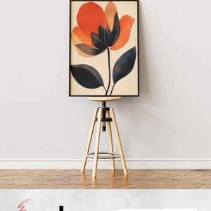 May include: A framed abstract art print featuring a large orange flower with black leaves on a cream background. The print is displayed on a wooden stool against a white wall.