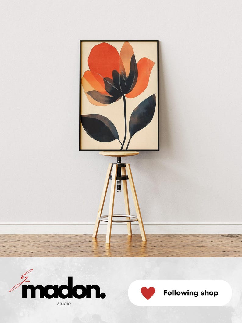 May include: A framed abstract art print featuring a large orange flower with black leaves on a cream background. The print is displayed on a wooden stool against a white wall.