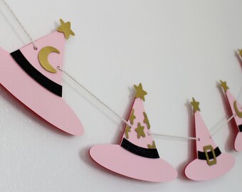 May include: A pink witch hat banner with gold accents. The hats have black bands and gold stars or moons. The banner is strung on a white string and is against a white wall.