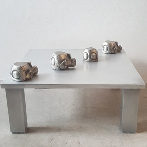 May include: Four silver-coloured fish figurines are arranged on a white square table with four legs. The figurines are all facing the same direction, with their tails pointing towards the edge of the table.