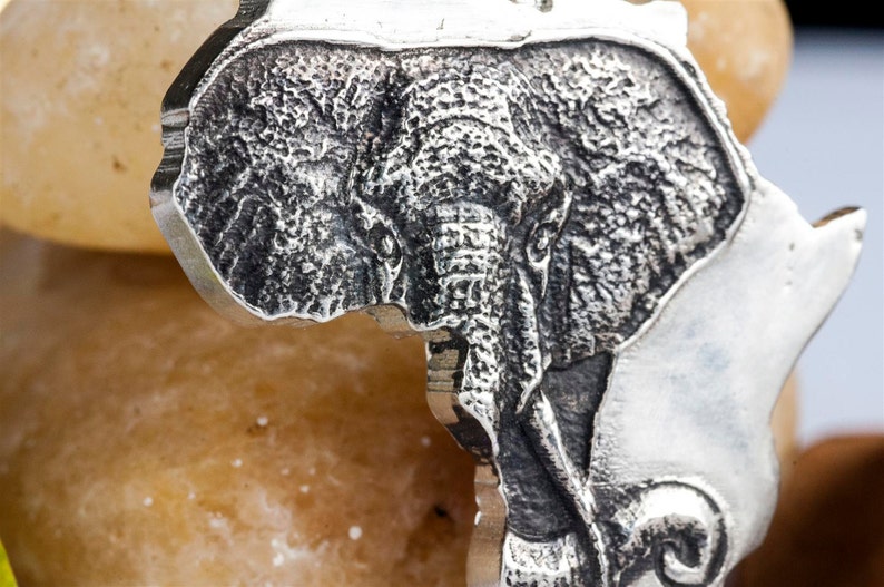 Op de afbeelding: Een zilveren hanger in de vorm van het Afrikaanse continent met een gegraveerd olifantenhoofd in het midden.