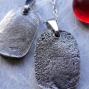 May include: Two silver fingerprint pendant necklaces. One pendant displays a detailed fingerprint pattern, while the other features a textured paw print. A red glass sphere is visible in the background. The pendants hang on delicate silver chains.