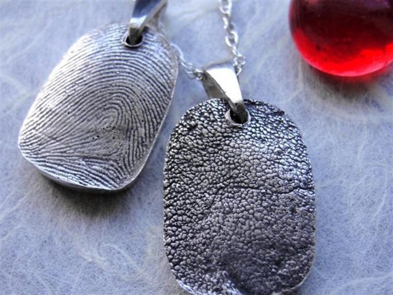 May include: Two silver fingerprint pendant necklaces. One pendant displays a detailed fingerprint pattern, while the other features a textured paw print. A red glass sphere is visible in the background. The pendants hang on delicate silver chains.