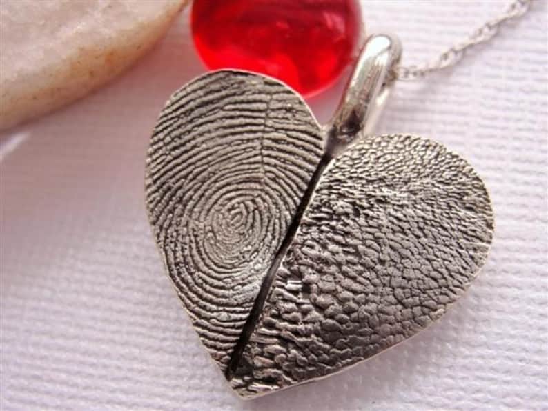 May include: A silver heart-shaped pendant necklace with two fingerprint impressions. The pendant is split in half, with each half featuring a different fingerprint.
