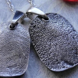 May include: Two silver pendant necklaces with unique textures. One pendant displays a detailed fingerprint pattern, while the other has a textured surface. Both pendants are rectangular with a loop for the chain.