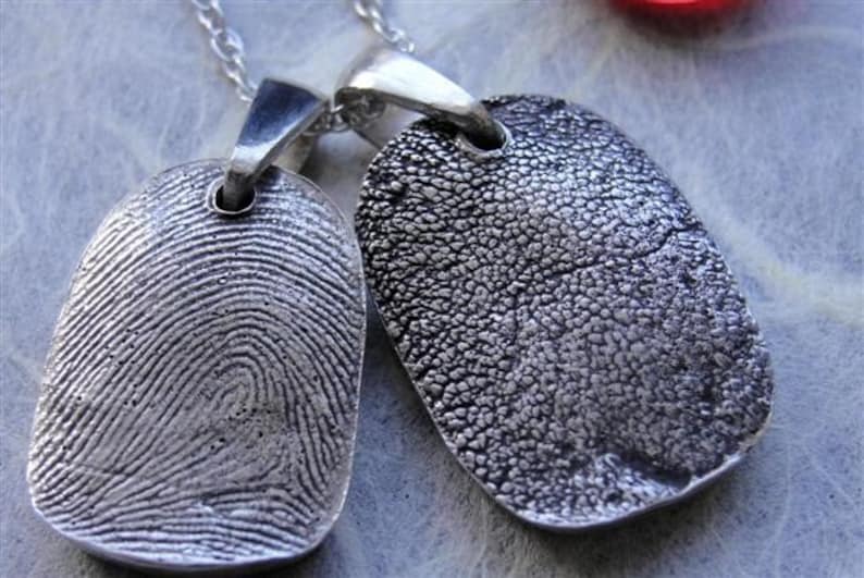 May include: Two silver pendant necklaces with unique textures. One pendant displays a detailed fingerprint pattern, while the other has a textured surface. Both pendants are rectangular with a loop for the chain.