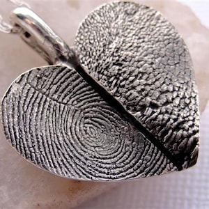 May include: A silver heart-shaped pendant with two fingerprint impressions. The pendant is split in half, with each half featuring a different fingerprint.