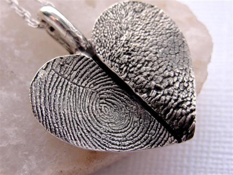 May include: A silver heart-shaped pendant with two fingerprint impressions. The pendant is split in half, with each half featuring a different fingerprint.