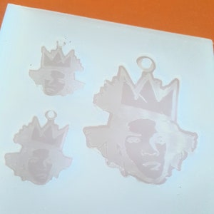 Puede incluir: Un molde de silicona rosa claro con tres dise&ntilde;os de caras con coronas. Los dise&ntilde;os son de diferentes tama&ntilde;os, con un lazo en la parte superior para colgar. Las caras tienen rasgos detallados y las coronas tienen un dise&ntilde;o dentado.