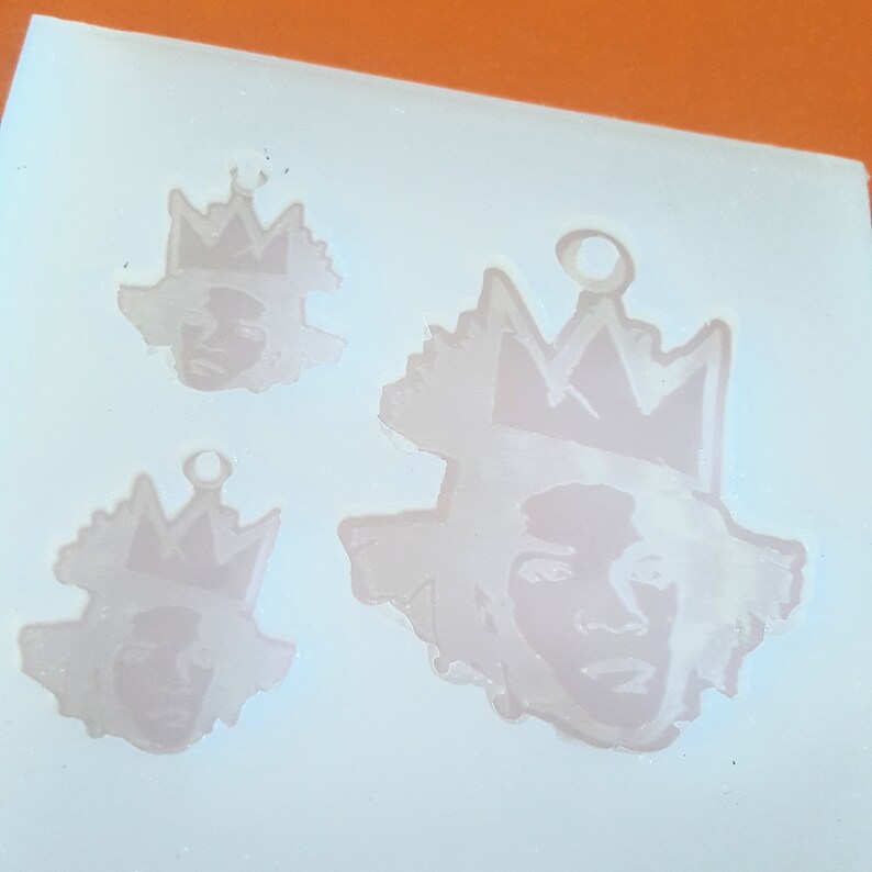 Puede incluir: Un molde de silicona rosa claro con tres dise&ntilde;os de caras con coronas. Los dise&ntilde;os son de diferentes tama&ntilde;os, con un lazo en la parte superior para colgar. Las caras tienen rasgos detallados y las coronas tienen un dise&ntilde;o dentado.