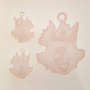 Puede incluir: Tres moldes de colgantes de resina rosa claro. Cada molde presenta una cara con una corona. Los moldes son de diferentes tama&ntilde;os, con un peque&ntilde;o lazo en la parte superior para colgar. El fondo es rosa claro.
