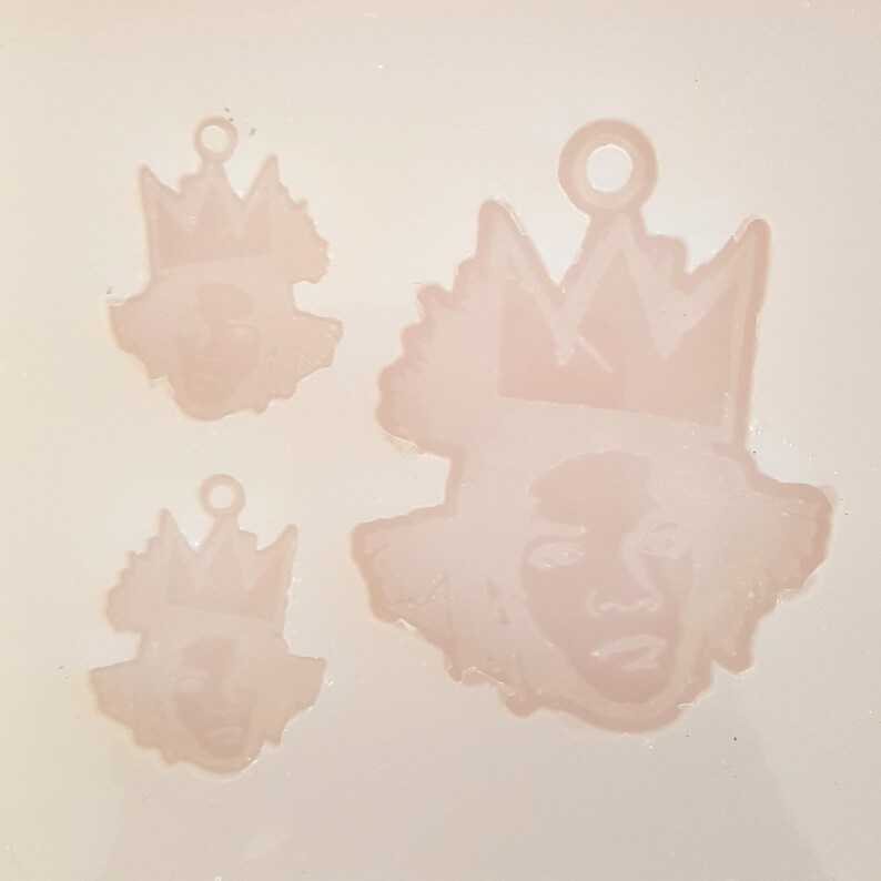 Puede incluir: Tres moldes de colgantes de resina rosa claro. Cada molde presenta una cara con una corona. Los moldes son de diferentes tama&ntilde;os, con un peque&ntilde;o lazo en la parte superior para colgar. El fondo es rosa claro.