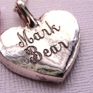 May include: Silver heart-shaped pendant with the inscription "Mark Bean" engraved on the front.