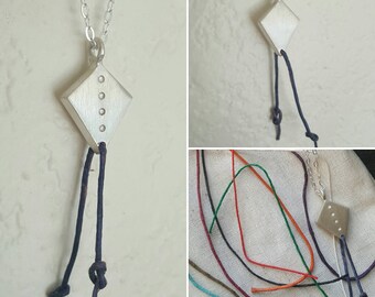 May include: A silver pendant shaped like a diamond with five small circles on the front. The pendant is attached to a silver chain and two black cords.