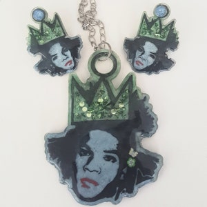Puede incluir: Un conjunto de collar y pendientes con un retrato con corona. El colgante y los pendientes tienen un fondo azul oscuro con un retrato de una persona con labios rojos y una corona verde. Los pendientes tienen una peque&ntilde;a gema azul en la parte superior.