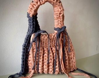 May include: A handmade, peach-colored tote bag with a braided handle and dark gray accents. The bag features a woven texture and is adorned with dark gray ties. The design is simple yet stylish, perfect for carrying everyday essentials.