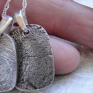 May include: Two silver pendant necklaces with unique fingerprint impressions. One pendant displays a detailed fingerprint pattern, while the other features a textured surface. Both are attached to delicate silver chains.