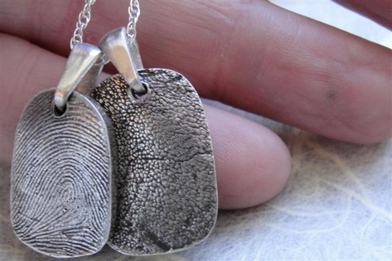 May include: Two silver pendant necklaces with unique fingerprint impressions. One pendant displays a detailed fingerprint pattern, while the other features a textured surface. Both are attached to delicate silver chains.