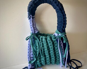 May include: A handmade tote bag in shades of blue and green. The bag features a braided navy blue handle and is constructed from thick, knitted fabric. The body of the bag is a textured, ribbed design with teal and lavender accents. Decorative ribbons are tied on the sides.
