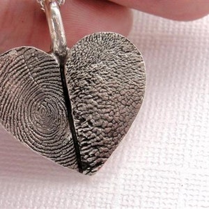 May include: A silver heart-shaped pendant with two fingerprint impressions. The pendant is on a silver chain.
