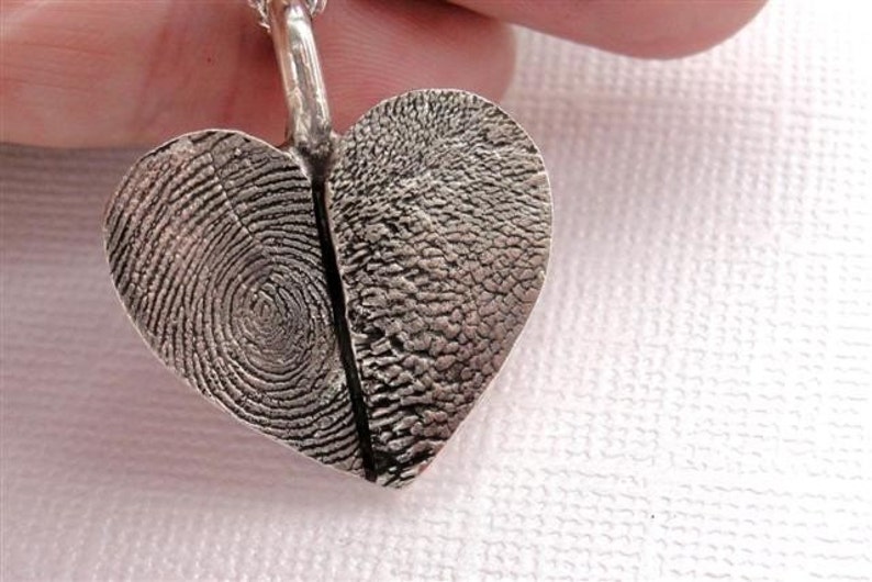 May include: A silver heart-shaped pendant with two fingerprint impressions. The pendant is on a silver chain.