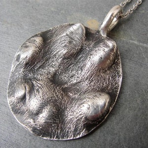 K&ouml;nnte beinhalten: Silberne Pfotenabdruck-Anh&auml;nger-Kette. Der Anh&auml;nger ist eine detaillierte, strukturierte Abformung einer Hundepfote. Die Kette ist aus Silber.