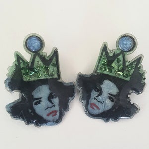 Puede incluir: Un par de pendientes de resina con un retrato coronado. Los retratos tienen labios rojos y est&aacute;n rematados con una corona verde con purpurina y una gema azul. Sobre un fondo blanco.