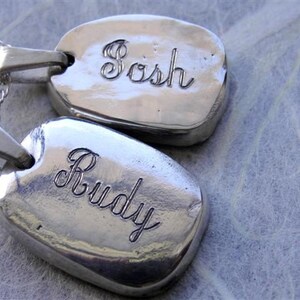 May include: Two silver-toned, oval-shaped pendants with the names "Sosh" and "Rudy" engraved in a cursive font. Each pendant hangs from a delicate silver chain. The background is a textured, light-colored surface.