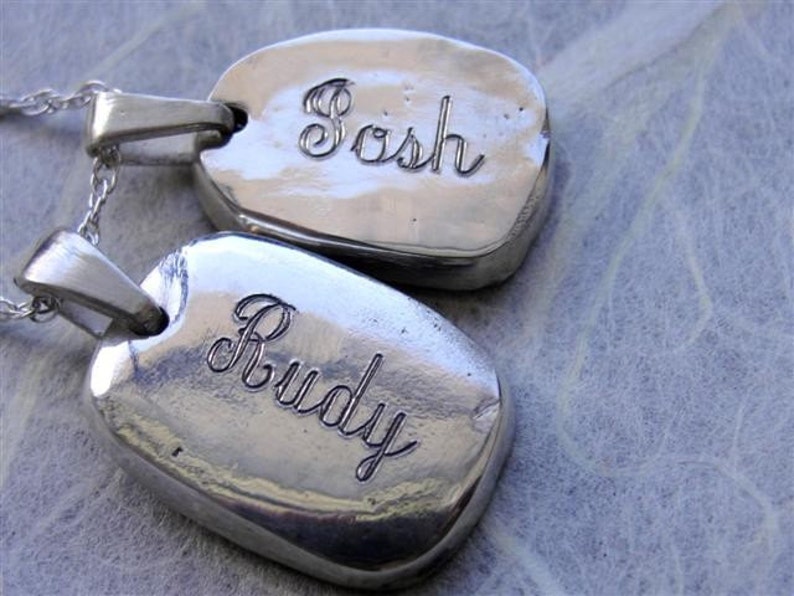 May include: Two silver-toned, oval-shaped pendants with the names "Sosh" and "Rudy" engraved in a cursive font. Each pendant hangs from a delicate silver chain. The background is a textured, light-colored surface.
