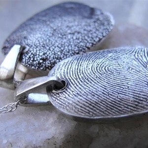 May include: Two silver fingerprint pendants on delicate chains. The pendants are oval-shaped with detailed fingerprint patterns. One pendant has a textured surface, while the other displays clear fingerprint ridges. The pendants are connected to silver chains.