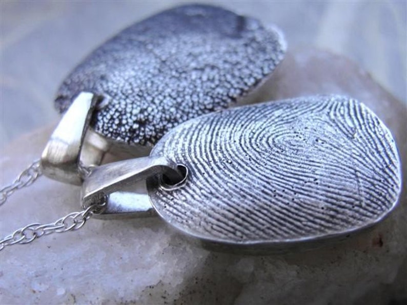 May include: Two silver fingerprint pendants on delicate chains. The pendants are oval-shaped with detailed fingerprint patterns. One pendant has a textured surface, while the other displays clear fingerprint ridges. The pendants are connected to silver chains.
