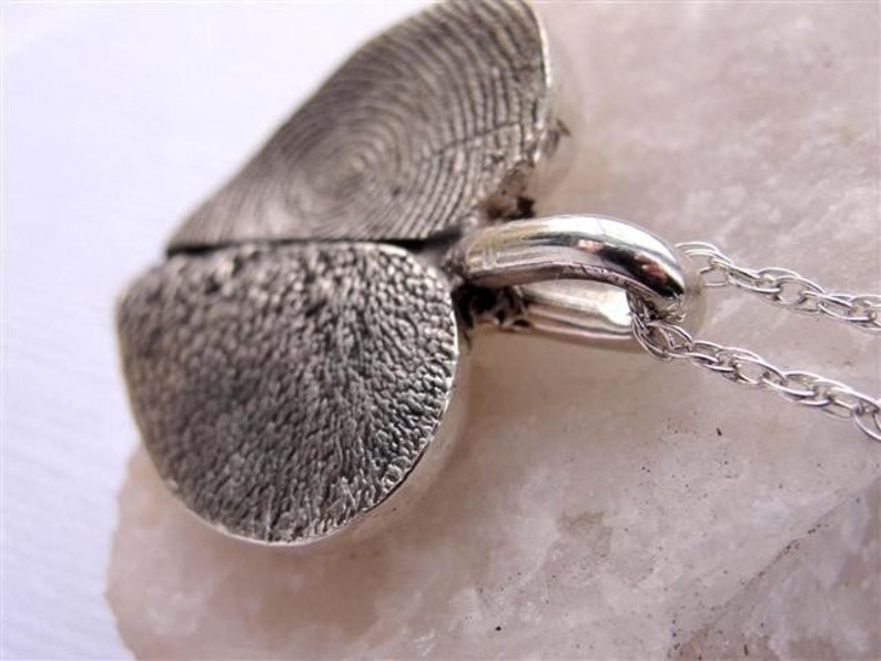 May include: A silver heart-shaped pendant necklace with a detailed textured design. The pendant is split in half and the two halves are connected by a silver chain.