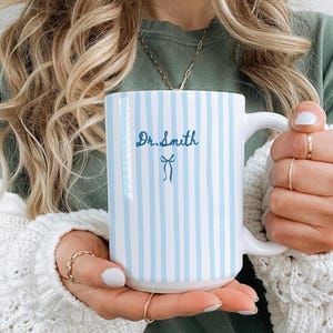 May include: A white ceramic mug with vertical light blue stripes. The mug features the name "Dr. Smith" in a script font, along with a small bow illustration. The mug has a large handle and is held by a person.