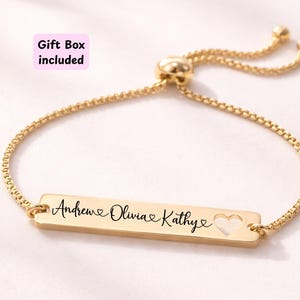 May include: A gold-coloured bracelet featuring a rectangular pendant engraved with the names "Andrew", "Olivia", and "Kathy", and a heart-shaped cutout. The bracelet has a delicate chain and a sliding clasp. The words "Gift Box included" are in a pink box.