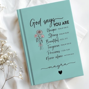 Puede incluir: Un diario turquesa con las palabras "Dios dice QUE ERES" y frases inspiradoras como "Único" y "Hermoso" en escritura negra. También se presenta una ilustración de un ramo floral y el nombre "mayra".
