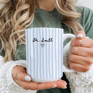 May include: A white ceramic mug with vertical light blue stripes. The mug features the text "Dr. Smith" in black script with a small heart symbol below. The mug has a large white handle.
